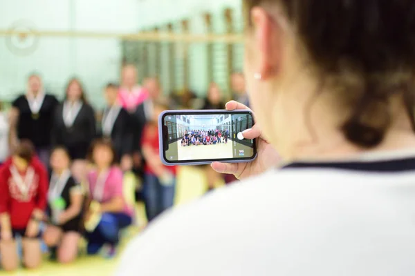 Kız telefonda kadın voleybol takımlarının ödüllerini fotoğraflıyor. Telefon ekranı odaklı ve arka planda kadınlar bulanık..