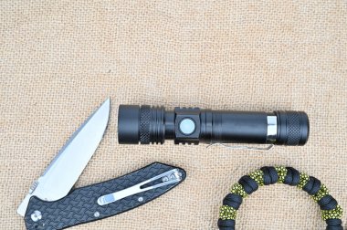 Hafifçe açılmış katlanmış bir bıçak, su geçirmez bir el feneri ve paraşüt bileziği. Her şey bir tuval yüzeyinde yatıyor..