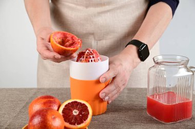 Kadın portakalın yarısını sağ elinde tutuyor ve meyve sıkacağı üzerine meyve suyu sıkıyor. Önünde portakallar var ve ikiye bölünmüş. İçinde sıkılmış meyve suyu olan bir sürahi var..