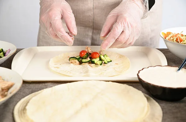 A woman in transparent gloves lays chopped vegetables, cucumbers and tomatoes on a round pita bread. The ingredients for shawarma are spread out around.