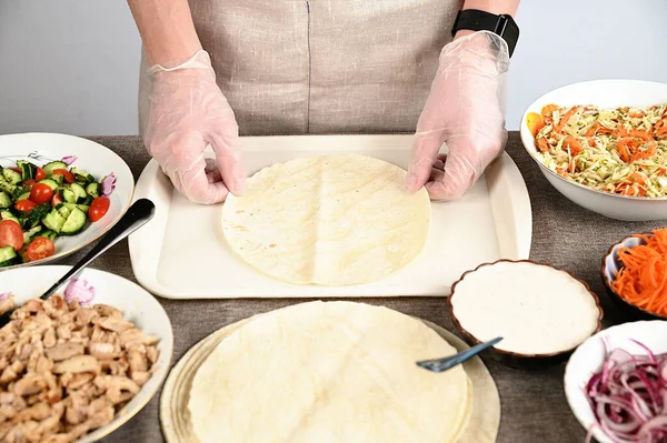 A woman in transparent gloves spreads a round pita bread on a white tray. There are ingredients for cooking kebab around.