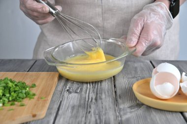 Bir kadın şeffaf bir tabakta omlet için yumurta çırpar. Yakınlarda doğranmış yeşil soğanlı ahşap bir tahta var..