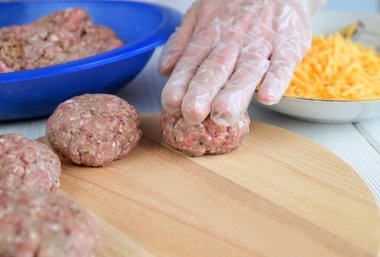 Şeffaf eldivenli bir kadın kıymalı pirzolaya bastırıp düz bir şekil veriyor. Tahta kesme tahtasındaki pirzolalar.