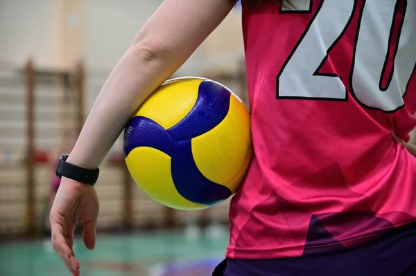 Bir voleybol oyuncusu, eli ve vücudu arasında voleybol topunu tutarken sırtı kameraya dönük durur. Numara futbol topunda görünüyor. Eline taşınabilir bir saat takılmış..