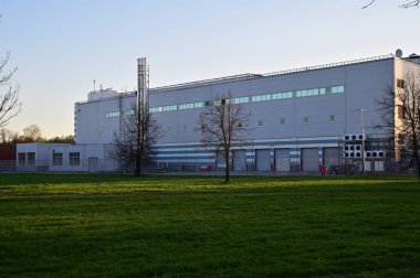 Küçük pencereli ve çitlerle çevrili büyük bir endüstri binası. Bina akşam güneşiyle aydınlanıyor..