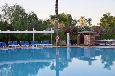 Deniz kıyısındaki bir otelde yüzme havuzu. Soyunma odası, güneşlik ve güneşlik. Ağaçtaki can simidi. Sabahın erken saatlerinde, güneş ağaçların tepelerinde parlıyor..