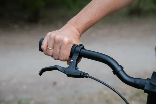 Bicycle handlebar with hand brake and a woman\'s hand holding the handle. A blurry park path in the background.