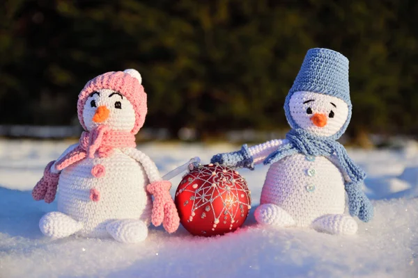 İki örülmüş kardan adam karda, parlak kış güneşinin altında duruyor. Aralarında bir Noel ağacı topu var. Arka planda daima yeşil bir ladin ormanı var..