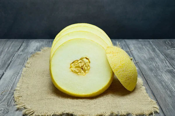 End view of a melon cut across into slices. The cap has been removed revealing a cut with seeds in the center. Melon lies on a wooden table. In the background is a gray cement wall.