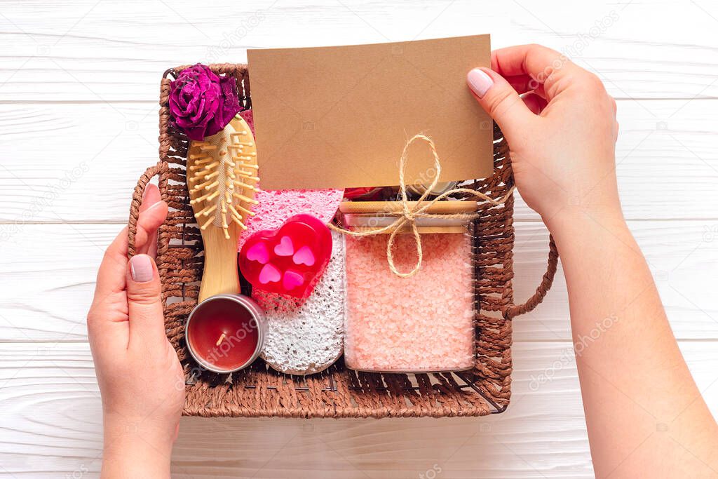 Female hand holds care box Set of eco - friendly cosmetics Bath salt, wooden comb, pumice stone, aroma candles, handmade soap, washing sponge on wooden background Gift for girlfriend, mother Top view