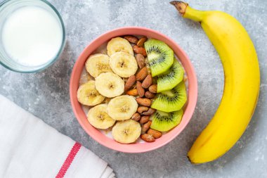 Muzlu yulaf ezmesi, badem fıstığı ve pembe kasede kivi dilimleri, bir bardak süt, gri beton arka planda peçete sağlıklı kahvaltı veya öğle yemeği..