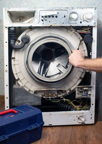 Washing machine repair. The front wall has been removed from the washing machine, the internal units and parts are visible. There is a box with a tool nearby. Repair of large household appliances