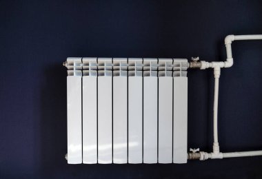 Beyaz ısıtma radyatörü mavi bir duvarın arkasında, pencereden gelen güneş ışığı ile aydınlatılıyor, ısıtma sistemine beyaz plastik borularla bağlanıyor.