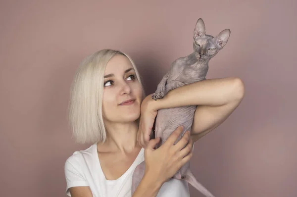 Güzel sarışın kadın gülümsüyor ve kollarında pembe arka planda kel bir Sphynx kedisi tutuyor.