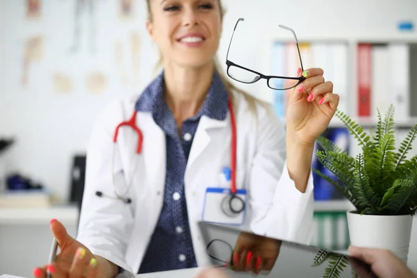 Mulher médico está sentado à mesa sorrindo e segurando óculos em suas mãos — Fotografia de Stock