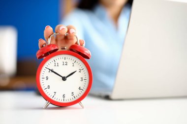 Womans hand lies on red alarm clock next to laptop. clipart