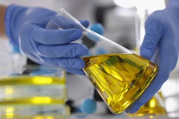Ruce v modrých rukavicích drží baňku s čirou zlatou tekutinou v laboratoři. — Stock fotografie