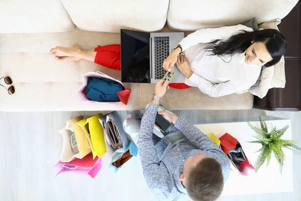 Frau sitzt mit Laptop auf Couch neben Mann und reicht ihre Bankkarte um Einkaufstüten — Stockfoto