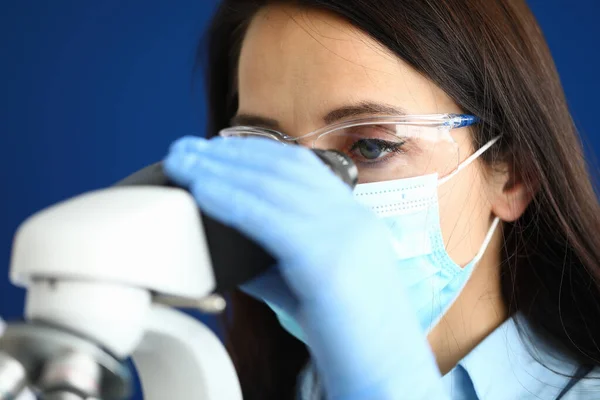 Kobieta w ochronnej masce medycznej w okularach i rękawiczkach patrzy przez mikroskop — Zdjęcie stockowe