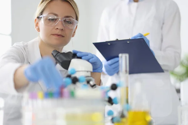 Forscher mit Brille nimmt Reagenzglas auf Tisch mit Mikroskop nahe Kollege schreibt die Ergebnisse auf. — Stockfoto