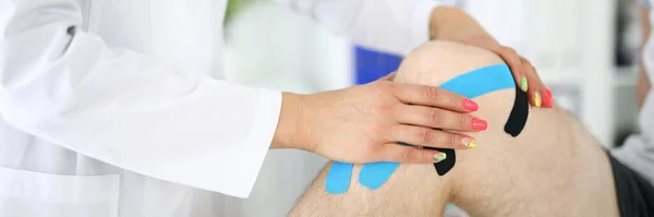 Traumatoloog lijmt blauwe en zwarte tape op patiënten pijnlijke knie in kliniek closeup — Stockfoto