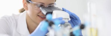 Lastik eldivenli ve koruyucu kimyasal gözlüklü bir kadın laboratuvar portresindeki mikroskoptan bakar.