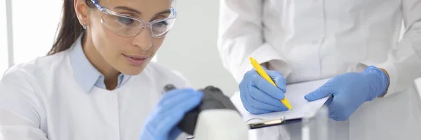 Junge Frau in weißer Uniform mit Chemikalienschutzbrille und Gummihandschuhen schaut ins Mikroskop — Stockfoto