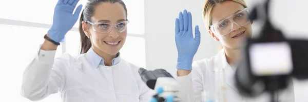 Fehér egyenruhás fiatal nők integetnek a kesztyűjükkel a kamera előtt a kémiai laborban. — Stock Fotó