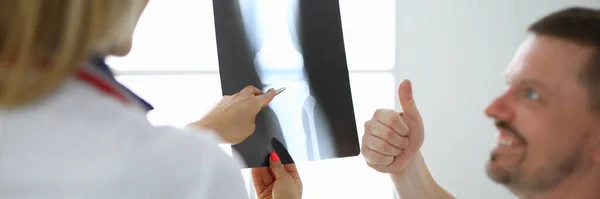 Médica mulher segura um raio-x da perna ao lado do paciente ele sorri e mostra polegares para cima — Fotografia de Stock
