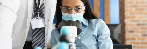 Жінка в захисній медичній масці і окулярах дивиться через мікроскоп поруч з колегою в білому пальто . — стокове фото