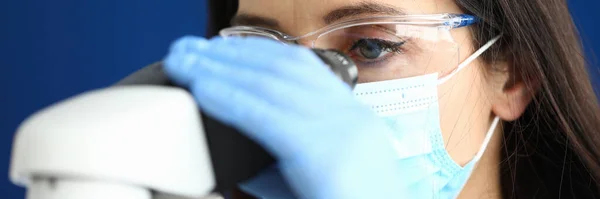 Kvinna i skyddande medicinsk mask med glasögon och handskar tittar genom ett mikroskop — Stockfoto