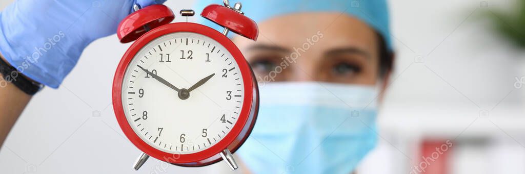 Doctor in medical protective mask holds red alarm clock in his hands