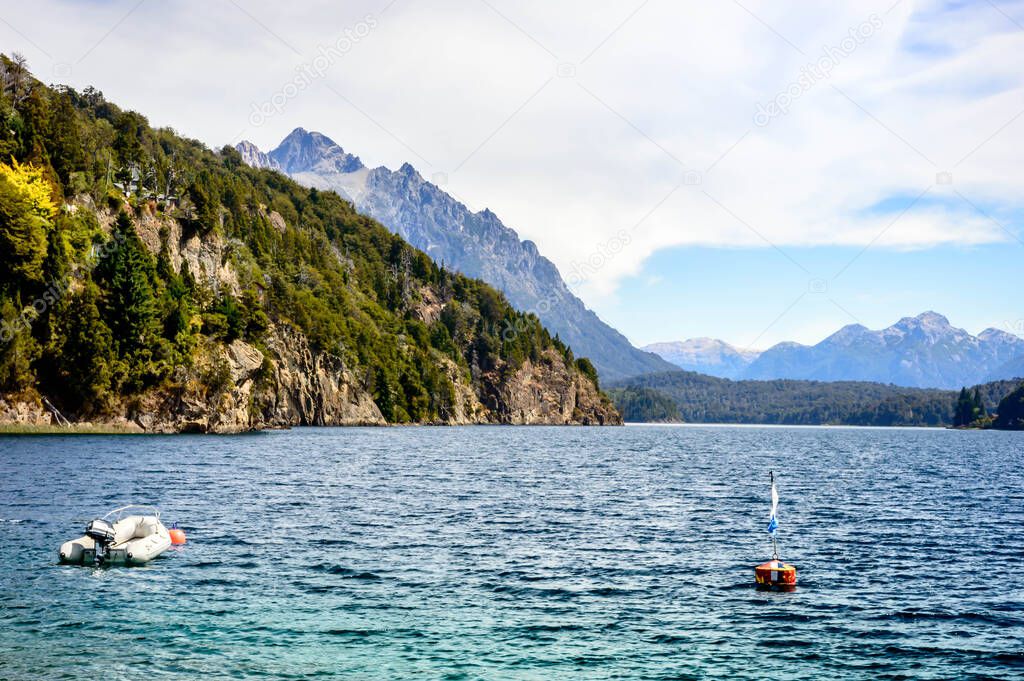 Lake shore with rocks, mountains and pine trees. A motorboat floating near a buoy. Summer in Bariloche.