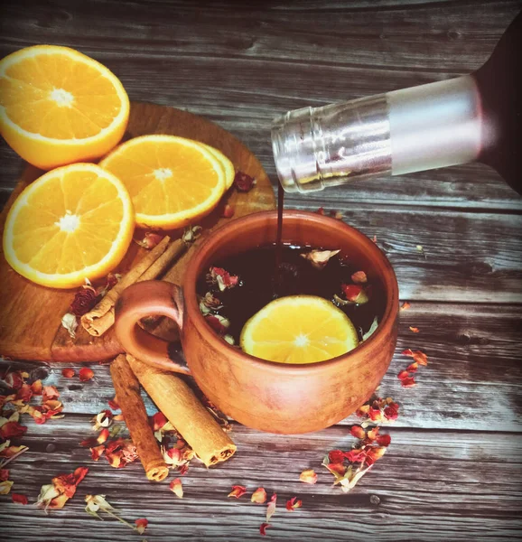 red mulled wine drink with the addition of cinnamon sticks, orange and fragrant dried rose petals on a wooden surface, wine is poured from the bottle in a small stream