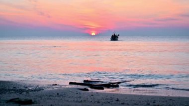 Güneş doğarken kumlu bir sahil manzaralı renkli bir video ve uzakta küçük bir balıkçı teknesi. Dramatik deniz gündoğumu, yumuşak pembe gökyüzü ve parlak dalgalar..