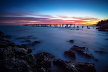 Gün batımından sonra uzun deniz manzarası ve kopmuş bir köprünün silueti.
