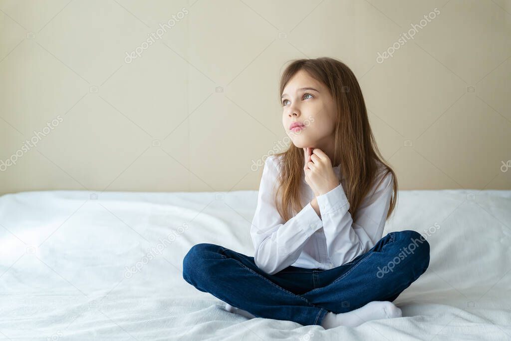 Close up shot of beautiful blonde caucasian little girl wearing jeans and a white shirt, sitting on a bed and looking calm and dreamy 