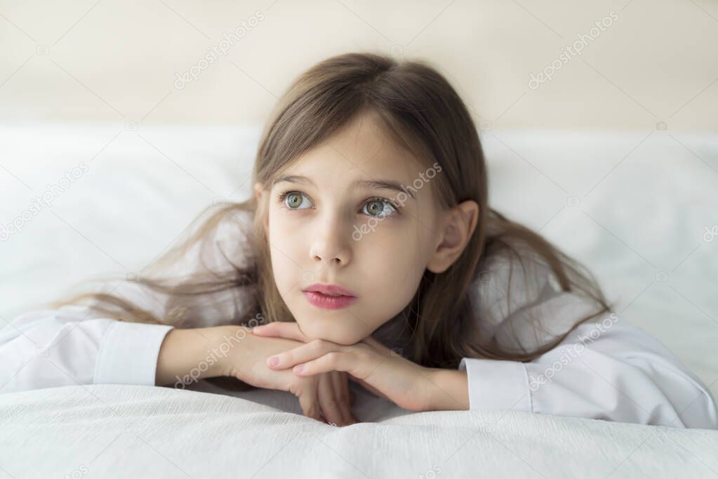 Close up shot of beautiful blonde caucasian little girl lying on a bed in a white shirt, looking calm and dreamy 