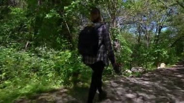Sırt çantalı ve kameralı bir kadın bahar yeşil ormanında dar bir yolda yürüyor.