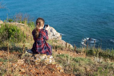 Turkuaz deniz sularıyla çevrili kayalık bir kıyı şeridinde oturmuş cep telefonuyla çalışan bir kızın arka görüntüsü.