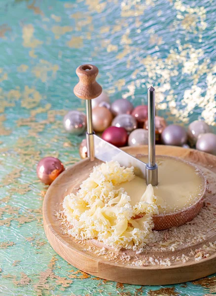 Fromage sur une planche ronde en bois et un couteau métallique spécial. Nouvel An, Noël, boules roses brillantes sur une surface verte. Photo De Stock