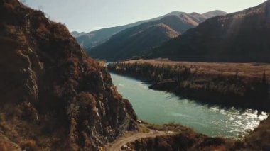 Sakin dağ nehri Katun 'un havadan görünüşü. Altai Bölgesi 'nin vahşi doğasının sonbahar manzarası: dağlar, ormanlar, tarlalar, kırsal bir yürüyüş yolu, güneş ışınları. Kamera uçuyor.