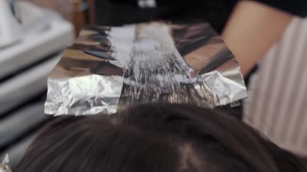 Gros plan sur le processus de mise en évidence des longs cheveux bruns d'une fille. Le coiffeur prépare les beaux cheveux forts de la fille pour mettre en évidence, peigner les cheveux, envelopper les cheveux humides dans du papier d'aluminium — Video