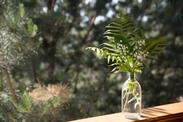 Fern leaves and lilies of the valley in a transparent vase-bottle stand on an old wooden windowsill against the background of a green garden. Spring and summer rural landscape.