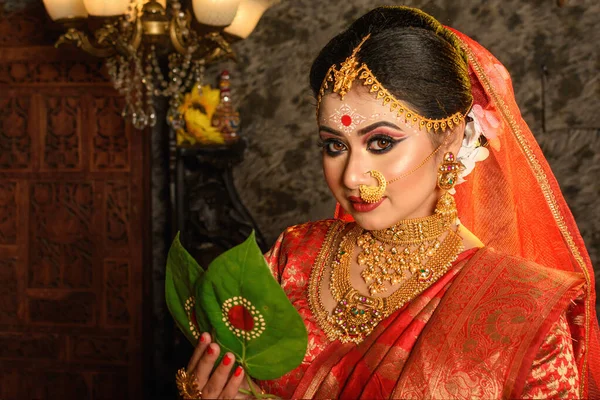 Portrait of very beautiful Indian bride holding betel leaf, Bengali bride in traditional wedding saree with makeup and heavy jewellery in studio lighting indoor