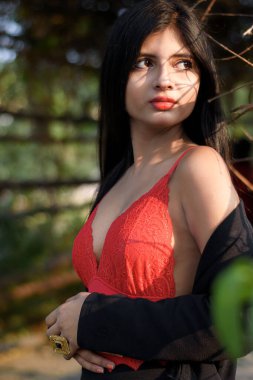 Güneşli bir günde, bulanık yeşil bir arkaplanda tropik yeşil yaprakların önünde poz veren kırmızı elbiseli çok güzel bir Hintli kadının portresi. Yaşam Tarzı ve Moda.