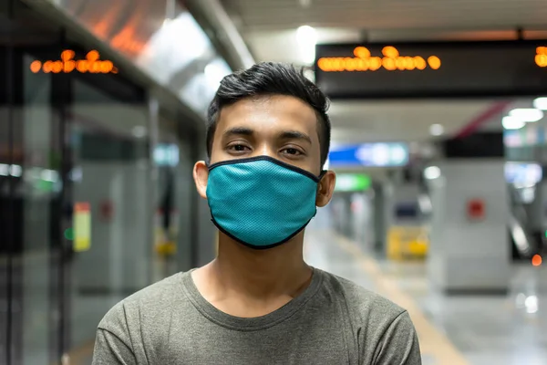 Tıbbi maskeli yakışıklı bir gencin tasviri sıradan bir tişörtle ayakta duruyor, kollarını kaldırıyor ve metro istasyonunda gülümseyerek kameraya bakıyor..