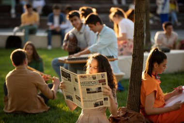 İstanbul, TURKEY - 01252021: Okul bahçesinde gazete okuyan kız. Diğer öğrenciler müzik yapıp sohbet ediyorlar. Güneşli bir günde kampüste keyifli zamanlar.