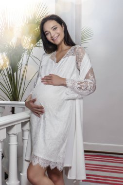 Genç, güzel, mutlu ve hamile bir kadın kameraya bakıyor ve gülümsüyor. Beyaz hamile elbisesi giyen esmer kadın elleri karnında poz veriyor. Mercek parlaması.