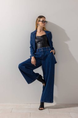 Büyük beden kot ceket ve deri bluzlu geniş paçalı kot giyen modacı kadın minimalist stüdyoda güven içinde poz veriyor. Modern eğilimler, temiz arkaplan ve alanı kopyala.